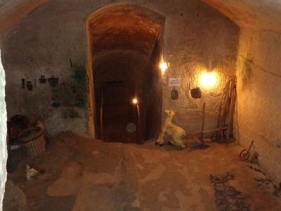 The image shows the interior of a rough-hewn stone cave dwelling with arched ceilings, featuring period-appropriate artifacts such as farming tools, a wooden cart, a ceramic jug, and a small statue of a goat, illuminated by warm artificial lightin
