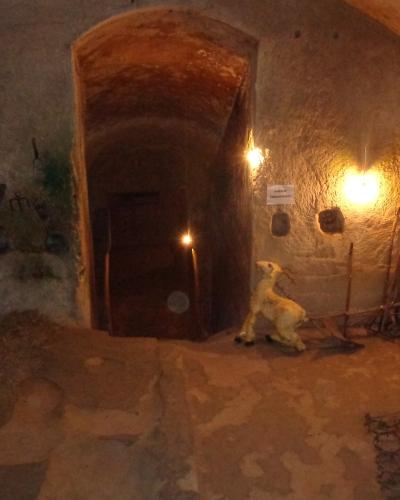 The image shows the interior of a rough-hewn stone cave dwelling with arched ceilings, featuring period-appropriate artifacts such as farming tools, a wooden cart, a ceramic jug, and a small statue of a goat, illuminated by warm artificial lightin