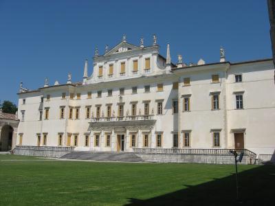 A large, white Baroque-style palace with a central pediment adorned with statues, multiple rows of windows with yellow shutters, and a grand staircase leading to the main entrance, set against a clear blue sky.