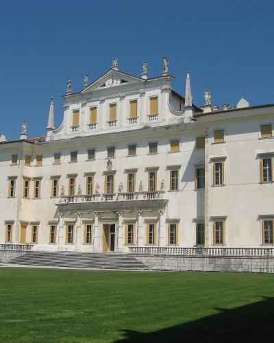 A large, white Baroque-style palace with a central pediment adorned with statues, multiple rows of windows with yellow shutters, and a grand staircase leading to the main entrance, set against a clear blue sky.