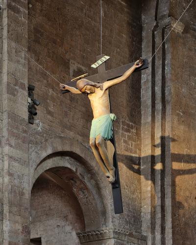A large crucifix statue of Jesus Christ suspended by cables against the stone facade of a medieval church, with an archway visible below.