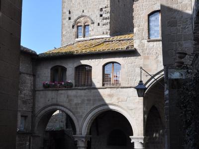 A stone courtyard with arched colonnades, a tall medieval tower with arched windows, and historic buildings under a clear blue sky; no visible signage or text identifying the location.
