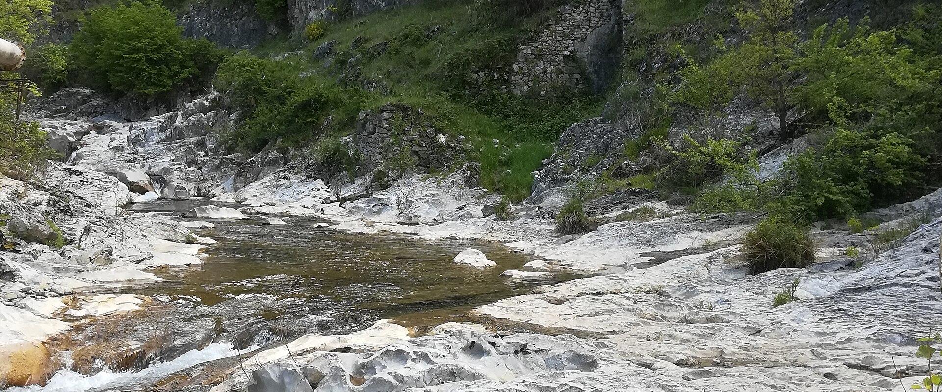 A shallow stream flows over a wide, white, heavily pitted and eroded limestone or travertine bedrock surface, with green forested hills rising in the background; no signage or human structures are visible.
