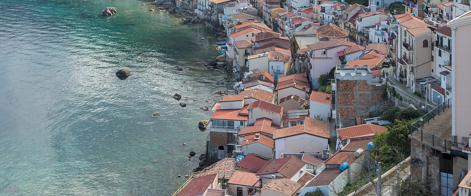 A high-angle view of a coastal village with terracotta-roofed buildings lining the shore, backed by green hills and a prominent castle on a cliff overlooking the sea.