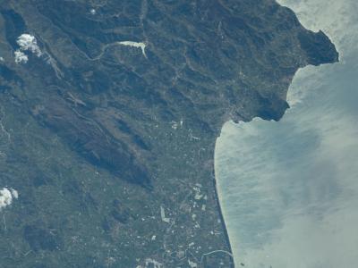 A high-altitude satellite or orbital photograph showing a rugged, mountainous coastline with a prominent peninsula jutting into the sea, a large inland lake (Lago Alento), and a flat coastal plain with agricultural fields and urban development nea
