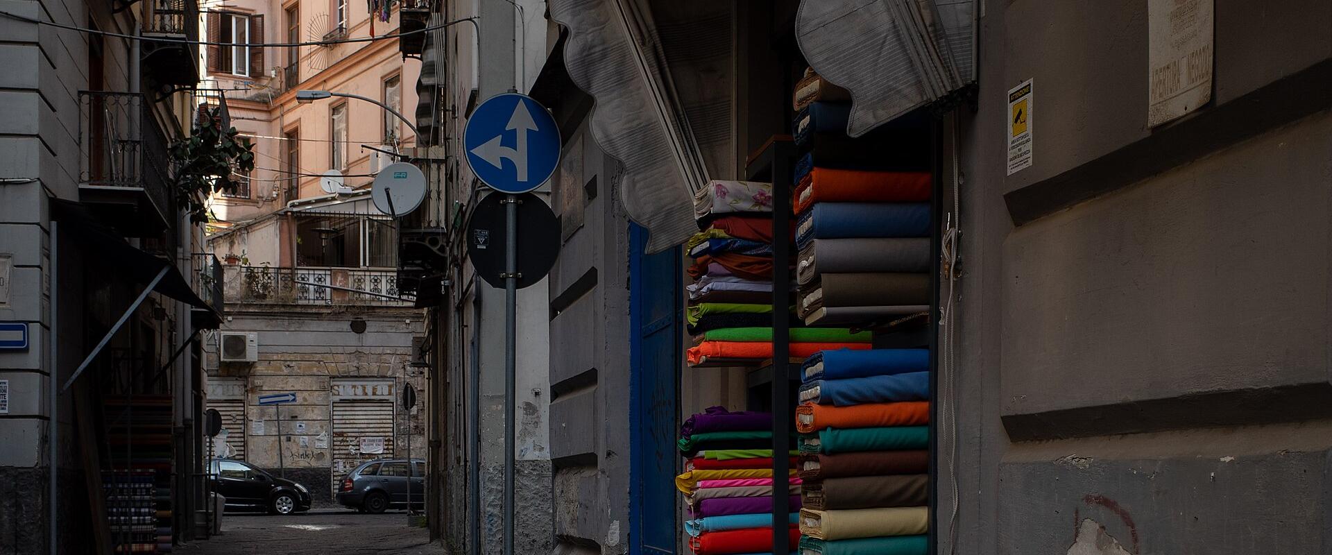 A narrow, cobblestone street in an urban setting with a fabric shop on the right displaying colorful bolts of cloth under a striped awning, and residential buildings with balconies and laundry in the background.