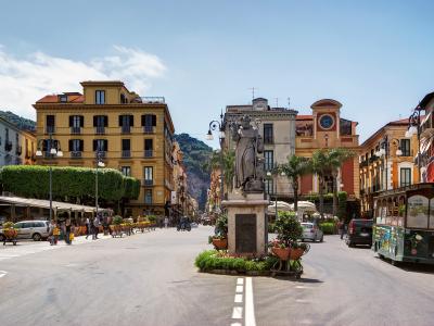A sunlit urban square in Italy featuring a central statue of a robed figure on a stone pedestal, flanked by multi-story buildings with shuttered windows, outdoor café seating, palm trees, and a green tourist tram on the right side.