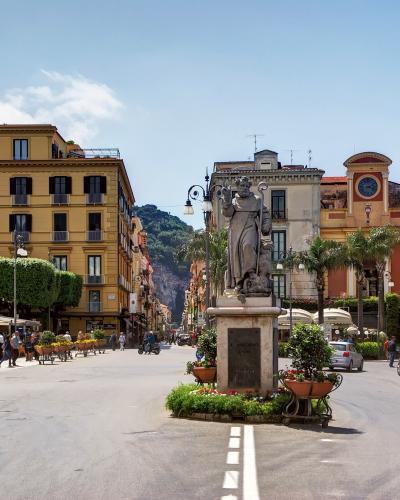 A sunlit urban square in Italy featuring a central statue of a robed figure on a stone pedestal, flanked by multi-story buildings with shuttered windows, outdoor café seating, palm trees, and a green tourist tram on the right side.