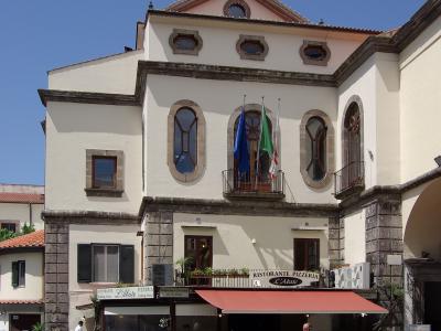 A cream-colored building with stone quoins and arched windows, housing a restaurant named "Ristorante Pizzeria L'Abate" on the ground floor, with flags displayed on a central balcony.