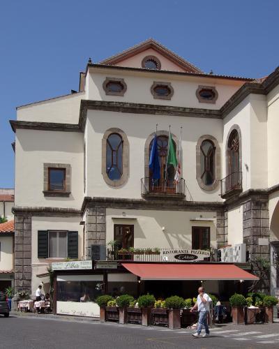 A cream-colored building with stone quoins and arched windows, housing a restaurant named "Ristorante Pizzeria L'Abate" on the ground floor, with flags displayed on a central balcony.