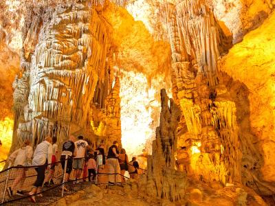 A group of tourists walking along a metal walkway inside a large, illuminated limestone cave filled with stalactites and stalagmites.