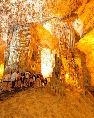 A group of tourists walking along a metal walkway inside a large, illuminated limestone cave filled with stalactites and stalagmites.