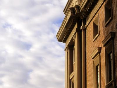 A low-angle architectural photograph showing the corner of a large, monumental building with rusticated brickwork, tall engaged columns, and a heavy classical cornice against a cloudy sky.