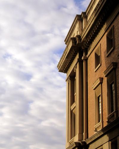 A low-angle architectural photograph showing the corner of a large, monumental building with rusticated brickwork, tall engaged columns, and a heavy classical cornice against a cloudy sky.