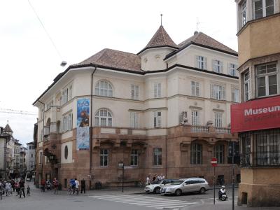 A street-level view of a large, light-colored building with a distinctive corner turret and terracotta roof, featuring a blue banner advertising "ÖTZI-MUSEUM" and a red sign reading "Museum im..." on an adjacent structure; people and vehicles are