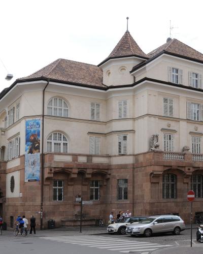 A street-level view of a large, light-colored building with a distinctive corner turret and terracotta roof, featuring a blue banner advertising "ÖTZI-MUSEUM" and a red sign reading "Museum im..." on an adjacent structure; people and vehicles are