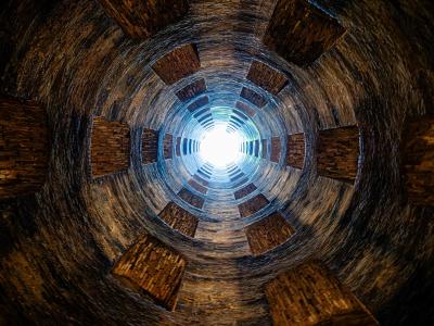 A low-angle, upward-looking photograph from inside a deep, cylindrical stone well, showing its spiraling double-helix staircase structure and the bright sky visible at the top opening.