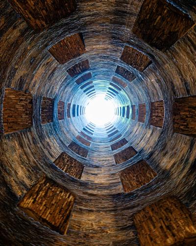 A low-angle, upward-looking photograph from inside a deep, cylindrical stone well, showing its spiraling double-helix staircase structure and the bright sky visible at the top opening.