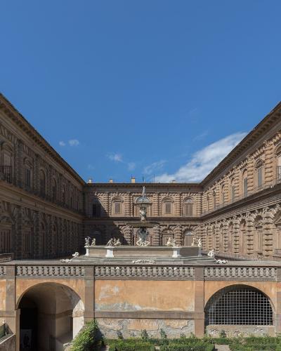 A view of a large Renaissance palace courtyard featuring rusticated stonework, a central fountain, and a balustrade in the foreground under a blue sky.