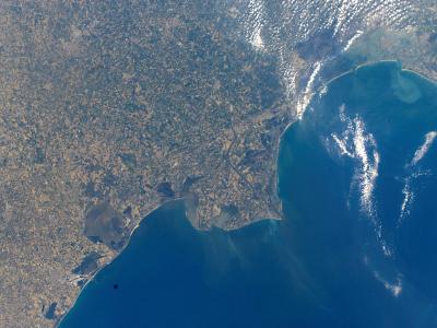 An orbital satellite photograph showing a coastal landmass meeting the sea, with visible sediment plumes in the water and cloud shadows on the surface.