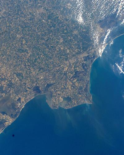 An orbital satellite photograph showing a coastal landmass meeting the sea, with visible sediment plumes in the water and cloud shadows on the surface.