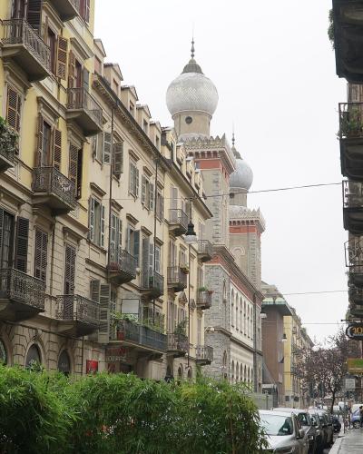 A street view in an urban European neighborhood featuring multi-story buildings with balconies and shutters, lined with parked cars and greenery, with a distinctive mosque featuring onion-shaped domes visible in the background.