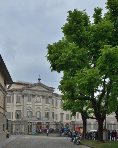 The image shows the neoclassical facade of a large building with columns, a pediment inscribed “ACCADEMIA CARRARA MDCCCX,” a clock, and an iron gate; people are gathered outside on a cobblestone street under a cloudy sky, with a large tree dominat