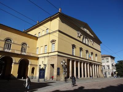 A low-angle exterior shot of a large, yellow neoclassical building with a colonnaded portico, arched windows, and a pediment featuring sculptural reliefs, set against a clear blue sky with overhead wires; people are walking and cycling in the fore