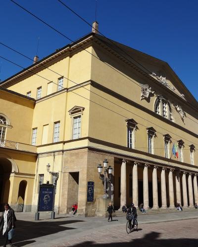 A low-angle exterior shot of a large, yellow neoclassical building with a colonnaded portico, arched windows, and a pediment featuring sculptural reliefs, set against a clear blue sky with overhead wires; people are walking and cycling in the fore