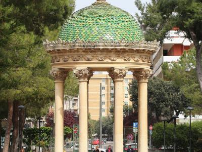 A classical-style rotunda with four columns and a green tiled dome topped by a cross, situated in a paved public park area with trees, benches, and pedestrians.