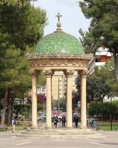 A classical-style rotunda with four columns and a green tiled dome topped by a cross, situated in a paved public park area with trees, benches, and pedestrians.