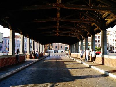 A view from inside a covered bridge looking down its length, showing a stone-paved walkway, brick parapets, stone columns, and a dark wooden truss roof structure with people walking and standing along the sides.