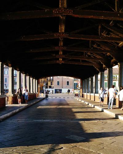 A view from inside a covered bridge looking down its length, showing a stone-paved walkway, brick parapets, stone columns, and a dark wooden truss roof structure with people walking and standing along the sides.