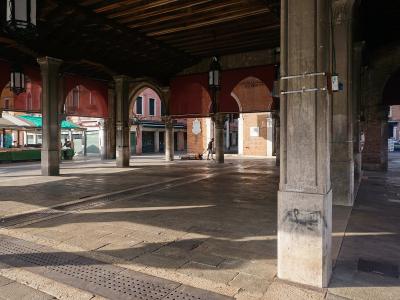 The image shows the interior of a covered market hall with stone columns, arches, and wooden ceiling beams, looking out onto a sunlit piazza with market stalls; signage on the brick wall reads "MERCATO DEL PESCE".