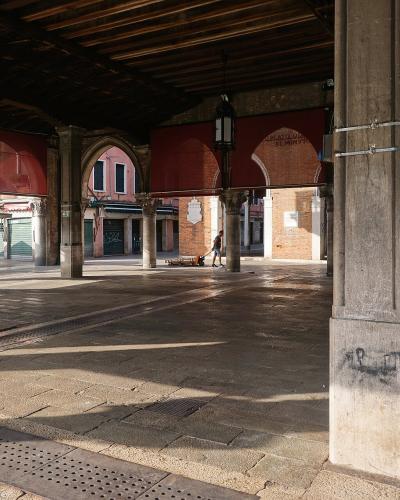 The image shows the interior of a covered market hall with stone columns, arches, and wooden ceiling beams, looking out onto a sunlit piazza with market stalls; signage on the brick wall reads "MERCATO DEL PESCE".