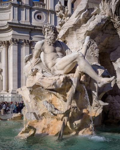 A close-up view of the Fontana dei Fiumi (Fountain of the Rivers) in Piazza Navona, Rome, featuring Gian Lorenzo Bernini's statue of the Nile river god with a rocky base and water flowing into the basin, with the facade of the Church of Sant'Agnes