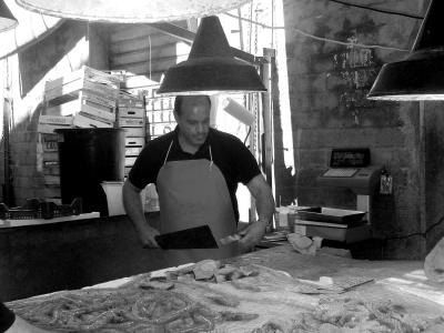 A black-and-white photograph showing a male fishmonger in an apron standing behind a counter laden with fresh seafood, including octopus and fish, under large industrial pendant lights in what appears to be an indoor market stall.