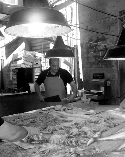 A black-and-white photograph showing a male fishmonger in an apron standing behind a counter laden with fresh seafood, including octopus and fish, under large industrial pendant lights in what appears to be an indoor market stall.