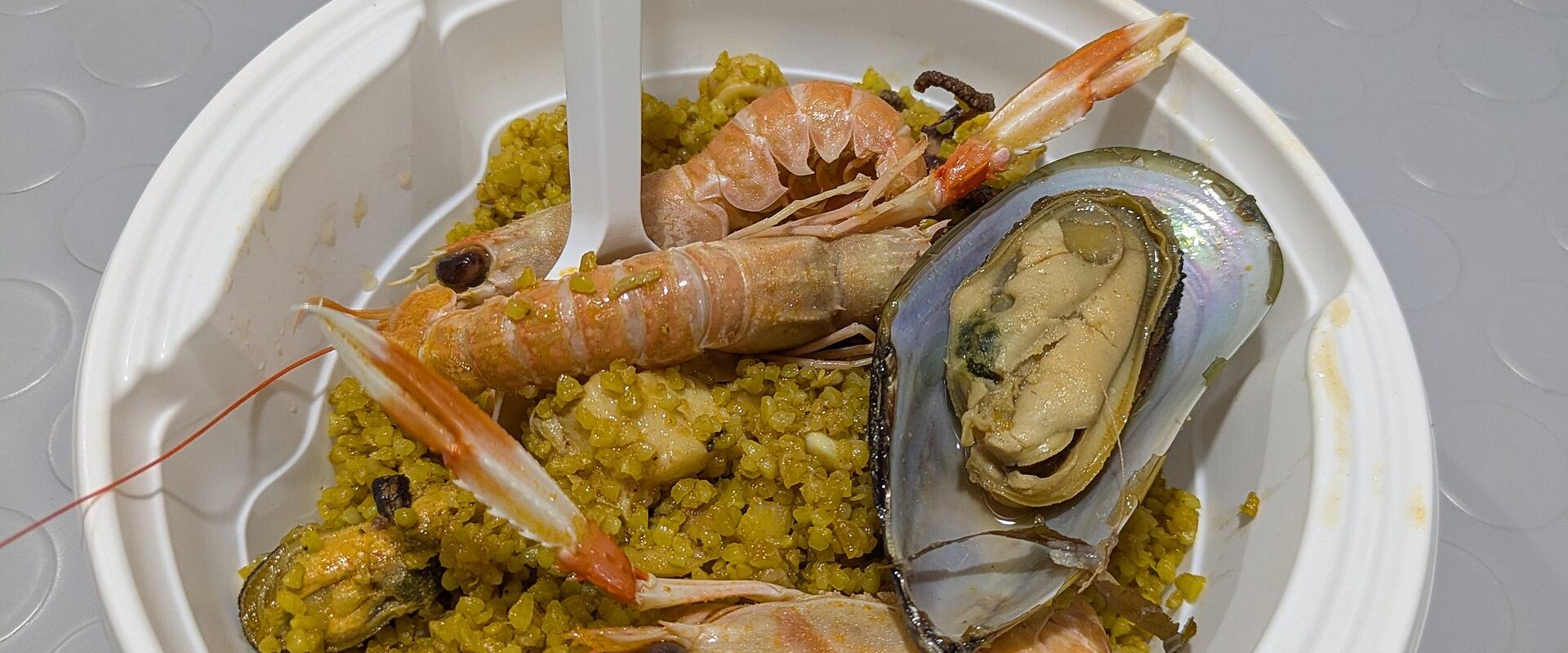 A close-up shot of a white disposable bowl containing yellow couscous topped with whole prawns, a mussel, and other seafood, with a white plastic fork inserted into the dish, resting on a grey textured table.