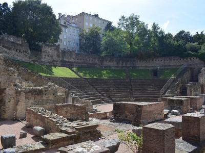 The image shows the ruins of an ancient Roman theatre with tiered stone seating, brick foundations, and modern metal railings, set on a hillside with residential buildings visible above.