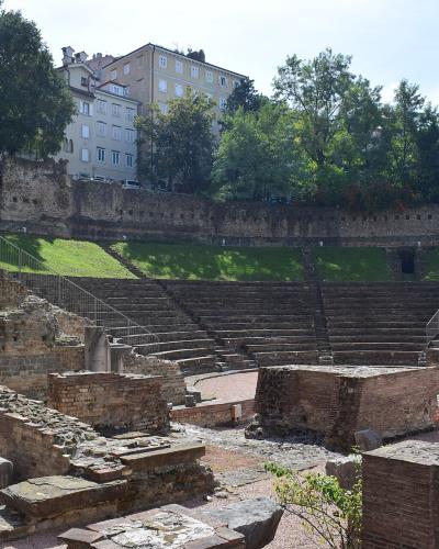 The image shows the ruins of an ancient Roman theatre with tiered stone seating, brick foundations, and modern metal railings, set on a hillside with residential buildings visible above.