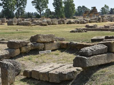 The image shows ancient stone ruins in a grassy field, with a small white sign reading "Tombe romane" (Roman tombs) placed among the blocks; in the background are more scattered stones and trees under a clear sky.