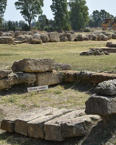 The image shows ancient stone ruins in a grassy field, with a small white sign reading "Tombe romane" (Roman tombs) placed among the blocks; in the background are more scattered stones and trees under a clear sky.