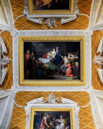 A view of an ornate interior ceiling and upper wall featuring three large, gilded-framed paintings set against a gold-leaf coffered ceiling with white stucco architectural details.