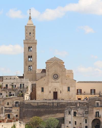 A wide-angle photograph of a large stone cathedral with a tall, square bell tower and a rose window, situated on a hilltop amidst other historic buildings under a blue sky.