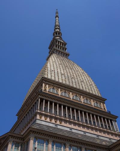 A low-angle photograph of a large, ornate building with a distinctive copper-clad dome and a tall, slender spire topped with a star, set against a clear blue sky.