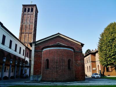 A red brick church with a rounded apse and a tall, square brick bell tower in the background, situated within a paved courtyard next to a white building with arches.