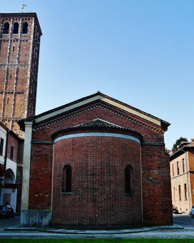 A red brick church with a rounded apse and a tall, square brick bell tower in the background, situated within a paved courtyard next to a white building with arches.