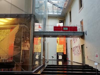 The image shows the interior atrium of a modern museum with glass walls, a metal bridge, and exhibition displays; visible signage includes "Back to BO" and "ABCDEF GHI LMNO PQR," indicating an Italian-language exhibit about Bologna.