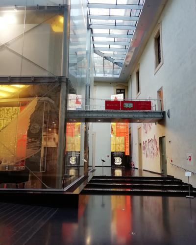 The image shows the interior atrium of a modern museum with glass walls, a metal bridge, and exhibition displays; visible signage includes "Back to BO" and "ABCDEF GHI LMNO PQR," indicating an Italian-language exhibit about Bologna.